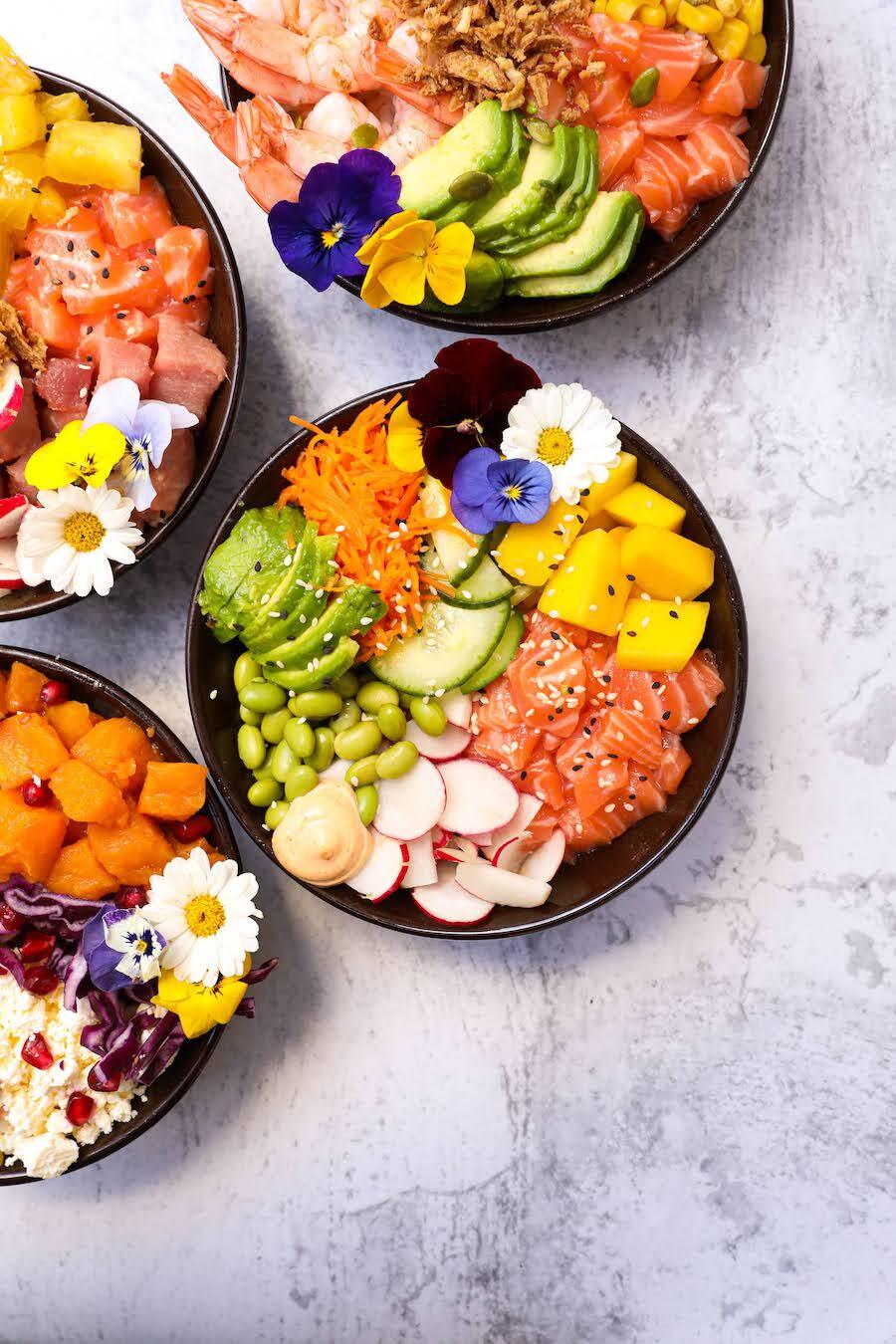 Assortiment de poke bowls colorés avec du saumon, des crevettes, des légumes frais, des fleurs comestibles, et des ingrédients exotiques, proposé par Mama's Secret